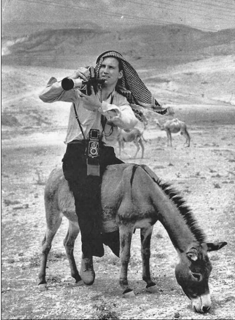 A man wearing a traditional Arabic head wrap perches on the back of a donkey, in a desert, while carrying a camera.