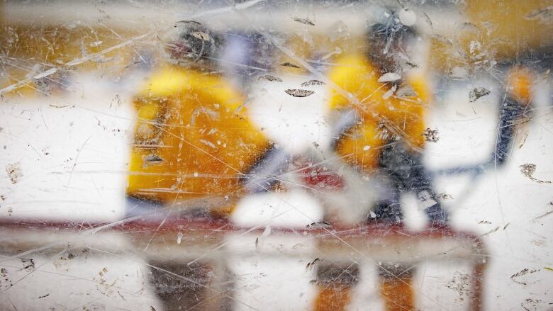 Two players in yellow jerseys are visible but blurry through the chipped and dirty arena glass.