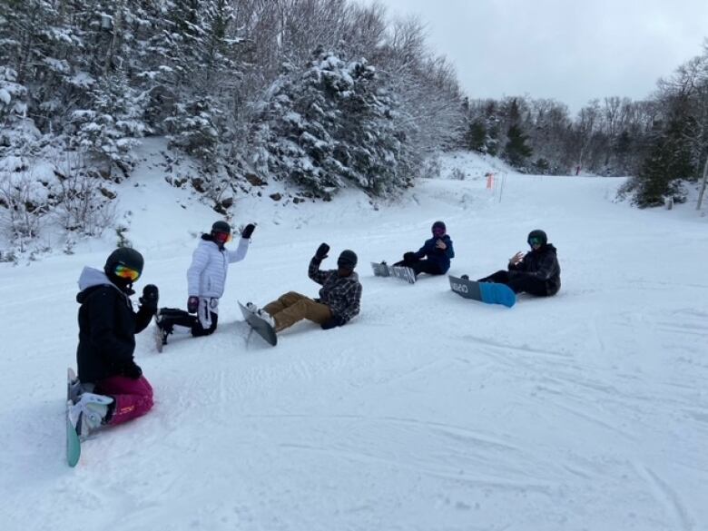 Five snowboarders wearing helmets and snow gear sit on a ski hill and wave. 