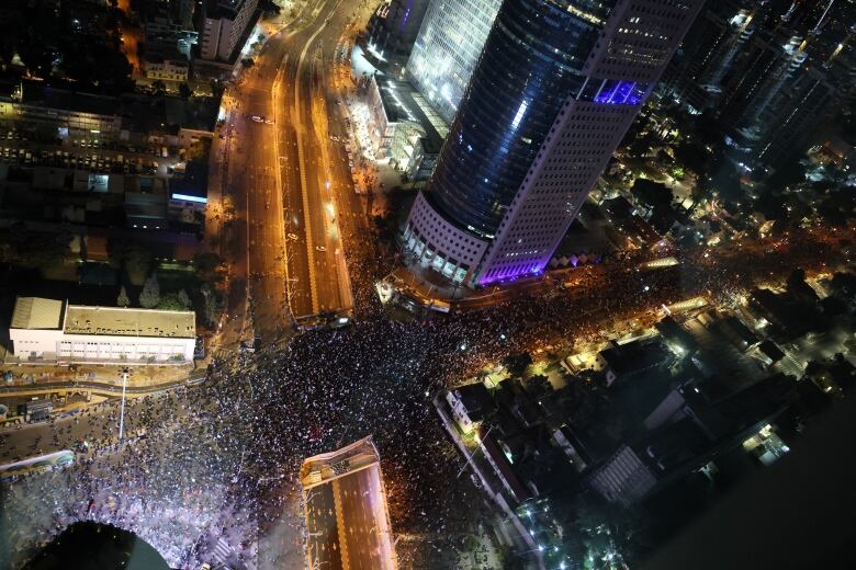 An aerial view at night of a large crowd of people filling intersecting streets between high-rise buildings.