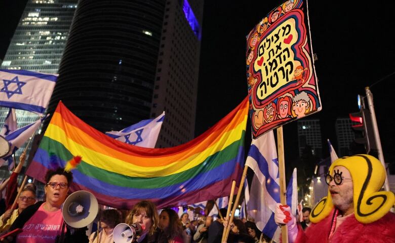 People hold up a long rainbow flag in front of a large crowd standing below skyscrapers at night.