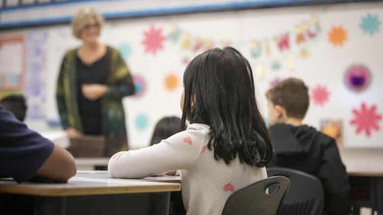 A teacher stands before children seated in a classroom. 