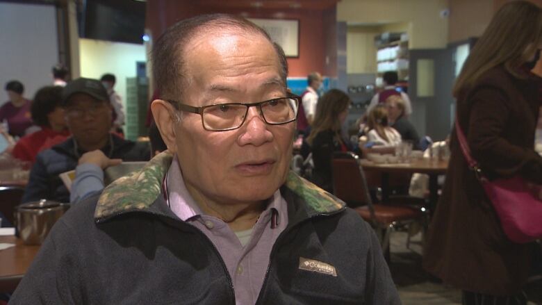 A man with glasses sits at a table with people in the background.