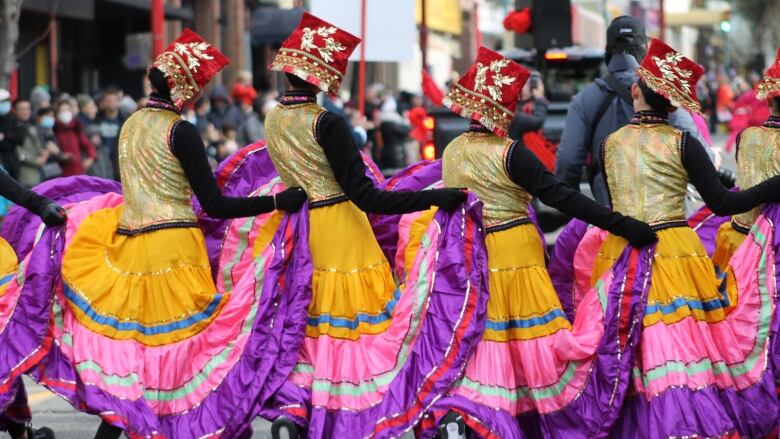 A row of dancers in fancy skirts on a city street, with numerous people lining up on the sidewalks in front of them.