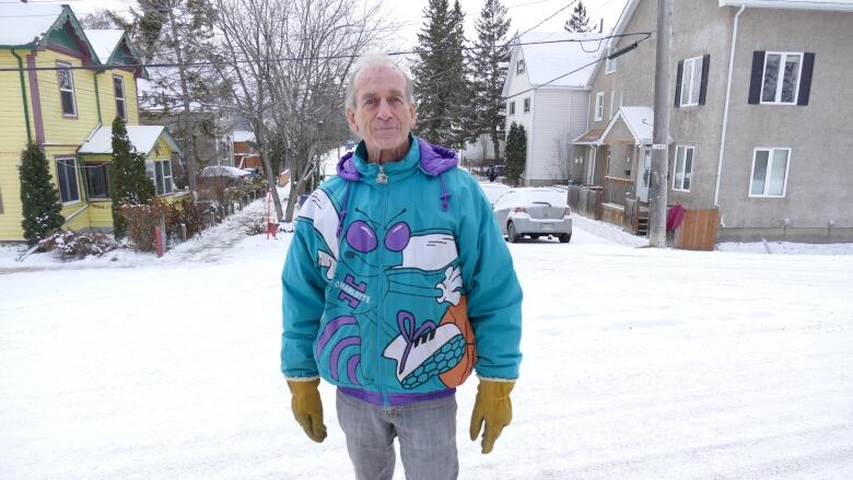 A man with white hair stands in a snowy street in front of two houses. He's wearing a teal and purple sweatshirt and jeans.