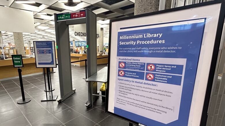 A walk-through metal detector stands in the background, to the left, while sign on a blue background in the foreground says 