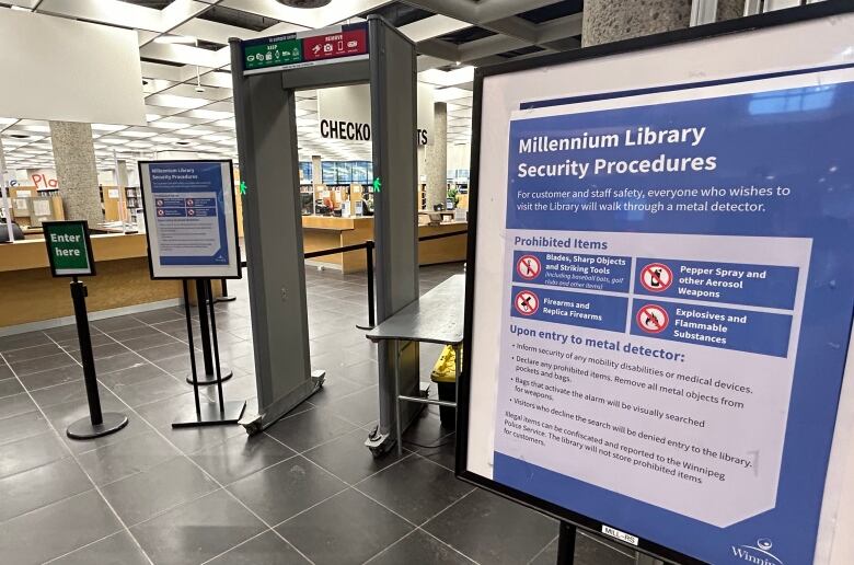 A walk-through metal detector stands in the background, to the left, while sign on a blue background in the foreground says 