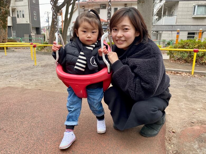 A two-year-old girl dressed in black pants, a black-and-white striped shirt and a black jacket sits in a red bucket swing at a park, while her mother, dressed all in black and smiling, crouches next to her for a portrait.