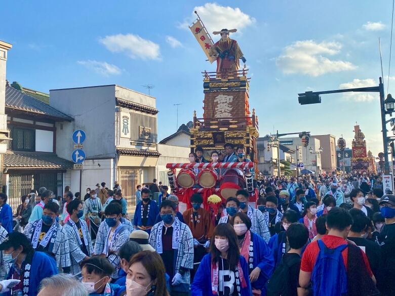 People dressed in white and blue fill the street, surrounding a large parade float that features a wooden tower and a figure holding a banner on top. People playing drums are at the base of the float.