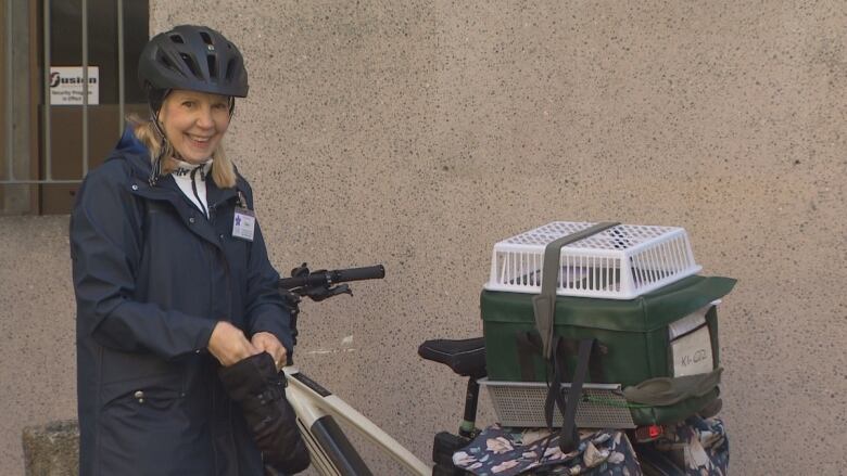 A blond woman in a bike helmet smiles as she stands next to a bike with a carrier fashioned on the back of it that contains meals.