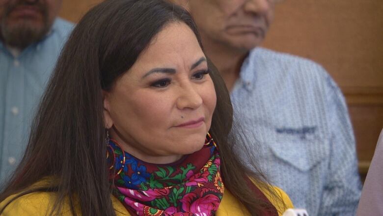 An Indigenous woman with long dark hair is wearing a red scarf with a flower design. The flowers are various colours: pink, blue, yellow and orange, with green for the stems. She is wearing a mustard-yellow blazer and earrings. Two men, each wearing button shirts tucked into their pants, stand in the background.