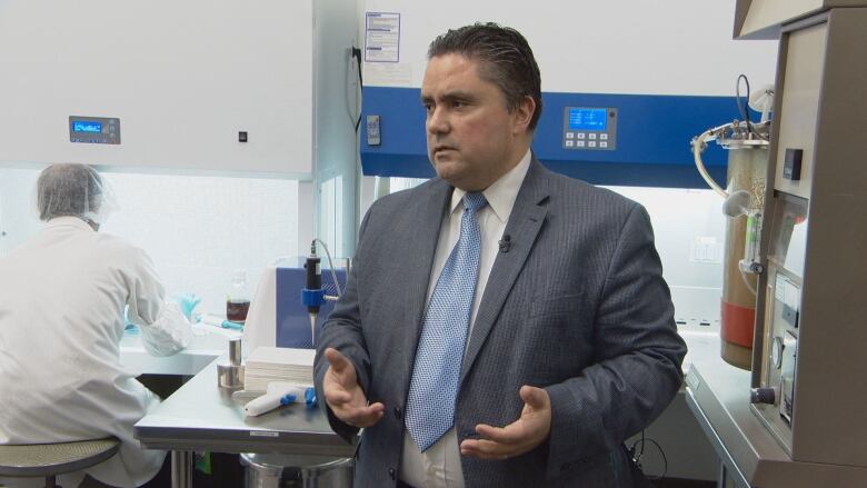 A man in a suit an tie stands in a laboratory next to a lab-coat-wearing staffer working under a fume hood and a vat churning a brown sludge.