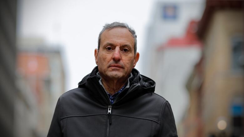 A white man with short grey hair looks directly at the camera with a neutral facial expression. He is outside with an urban street in the background and he is wearing a black zip-up jacket.