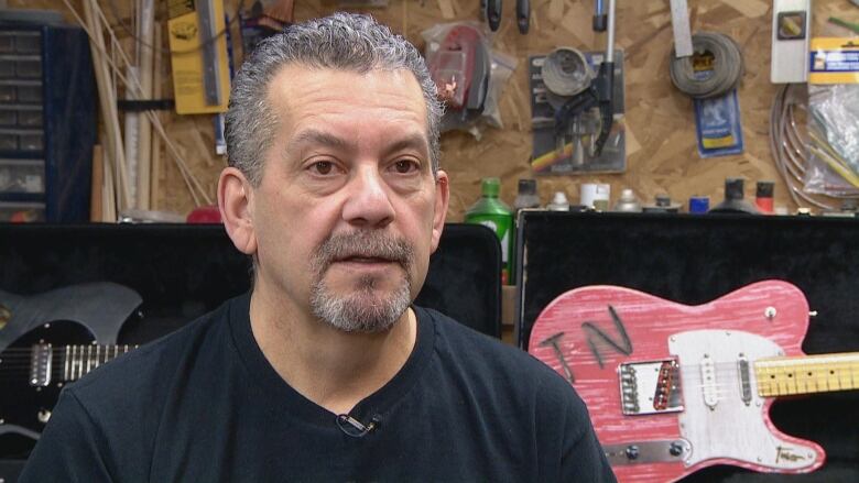 A man with a goatee and grey, short hair sits wearing a black T-shirt. Behind him is a pink custom guitar in the shape of a telecaster but with the letters 