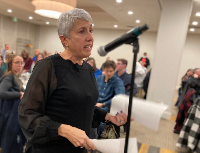 A woman with short white hair speaks into a microphone, crowd in the background