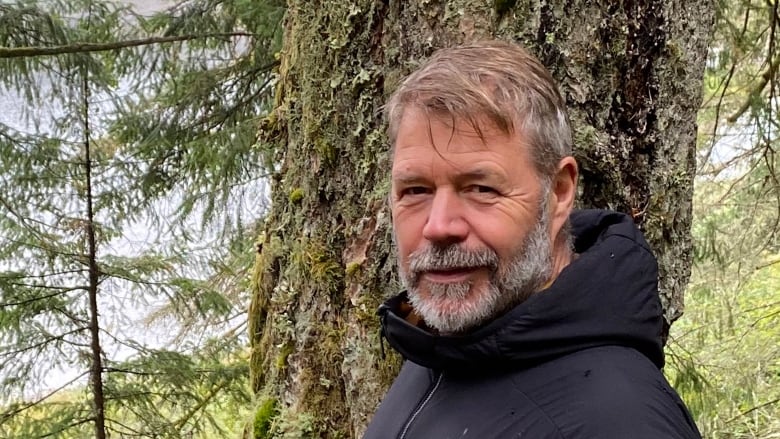 A man with a black raincoat and grey beard stands in the forest.