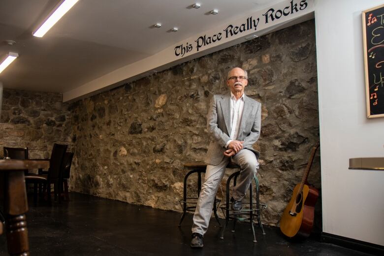 An older man in a suit with a white moustache sits on a stool with a guitar beside him.