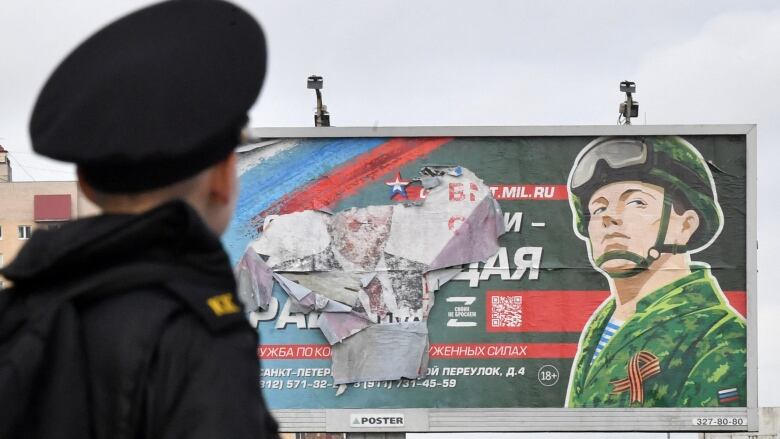 The back of the head of a young cadet looking at a weather-beaten billboard advertising army service in Russia. 