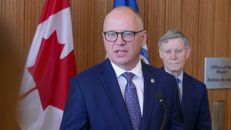 A man in a suit speaks at a podium. Behind him, another man in a suit listens, standing next to a Canadian flag.