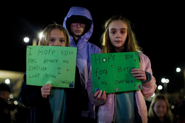 Three children stand together, two holding signs that contian Get Well messages for their tearcher.  