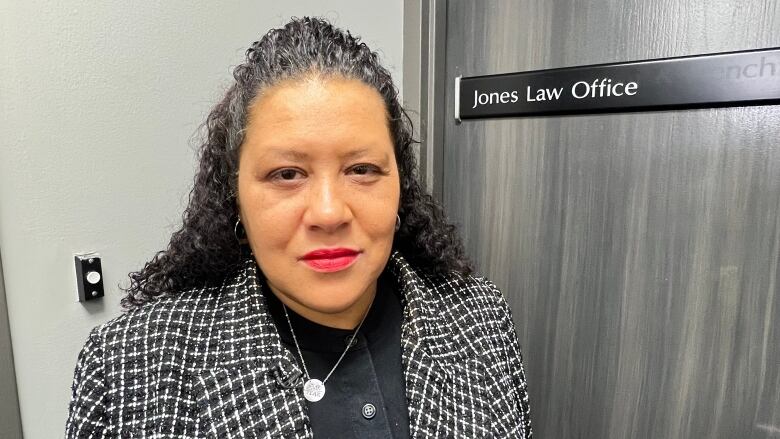 Zilla Jones is standing in front of a door with a name plate that says Jones Law Office. Part of her hair is pulled back. She is wearing a black and white checkered blazer and black dress.