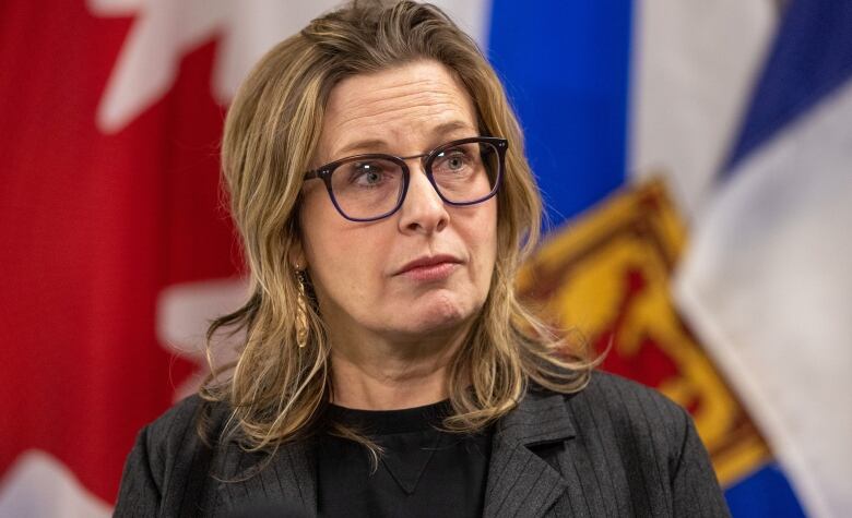 A woman with shoulder-length hair and glasses stands in front of the Canadian and Nova Scotia flags.