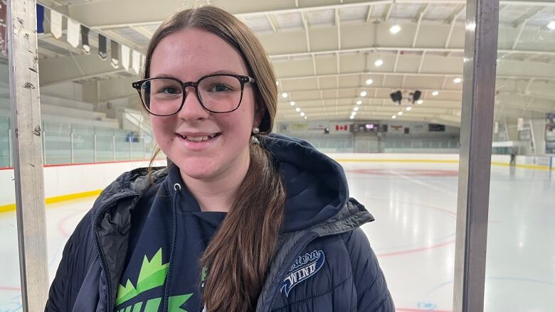 A girl wearing a Canada Games hoodie stands in front of an ice rink. 