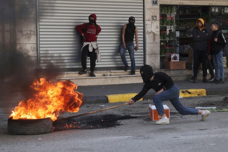 A person with a black mask on their head stokes a fire atop a tyre in the street.