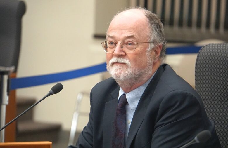 A man with a white and grey moustache, wearing a suit, sits at a microphone.