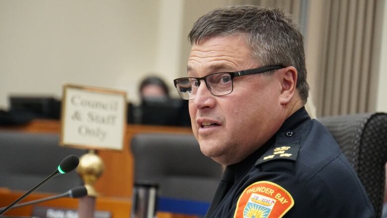 A uniformed police officer wearing glasses speaks at a microphone.