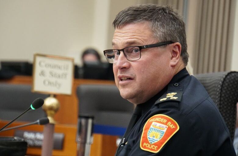 A uniformed police officer wearing glasses speaks at a microphone.