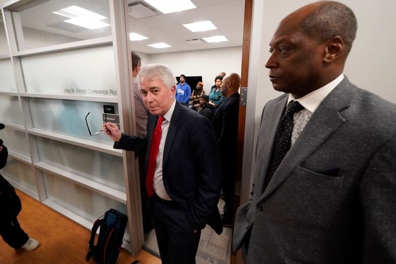 A man with grey hair walks out of a meeting room, as others look on. 