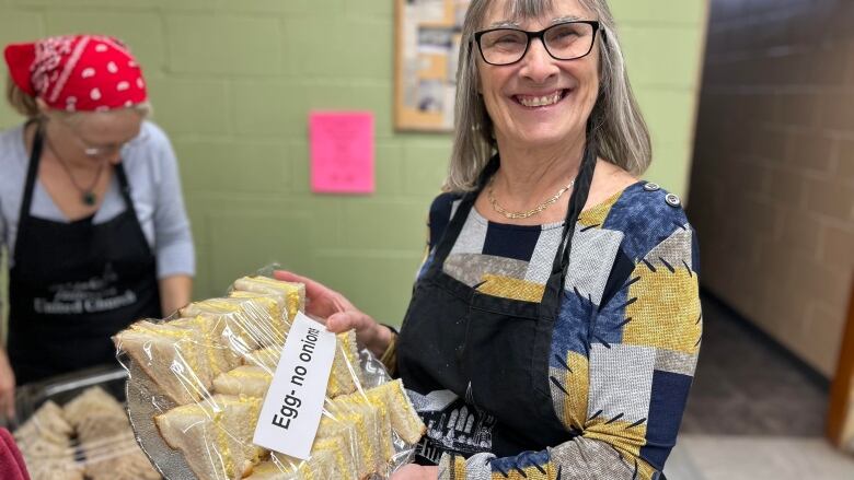 A woman in an apron holds a tray of sandwiches wrapped in plastic with a tag that reads 