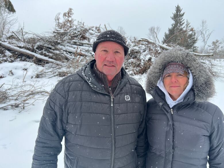 A man and woman in black winter coats stare through heavy snow toward the camera. The pile of tree trunks behind them is covered by snow.