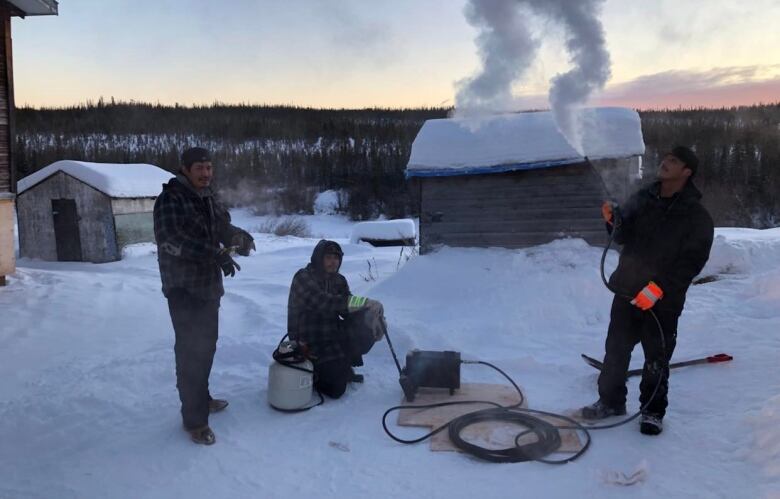 Three men outside in the snow. The one on the left is standing, the one in the middle is sitting next to a fuel tank, the one on the right is holding a wand that's flowing steam into the air.