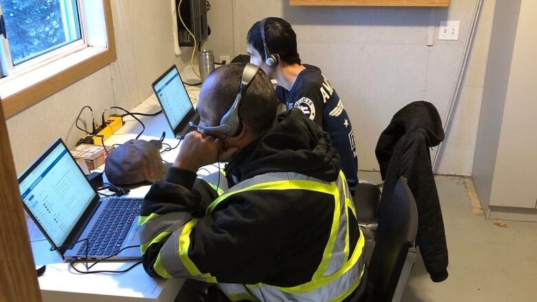 Two men seated infront of laptops, with headphones on, in a small room. 