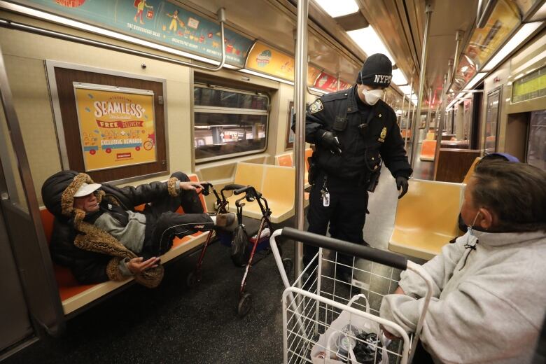 A police officer stands in a train carriage, talking to two people. One is lying down and the other is sitting.