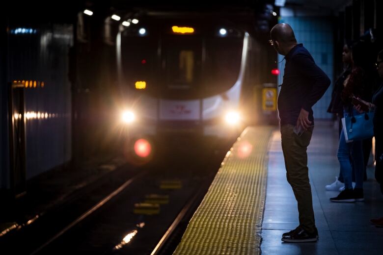 A dimly-lit image of a person on a subway platform looking towards an oncoming train.