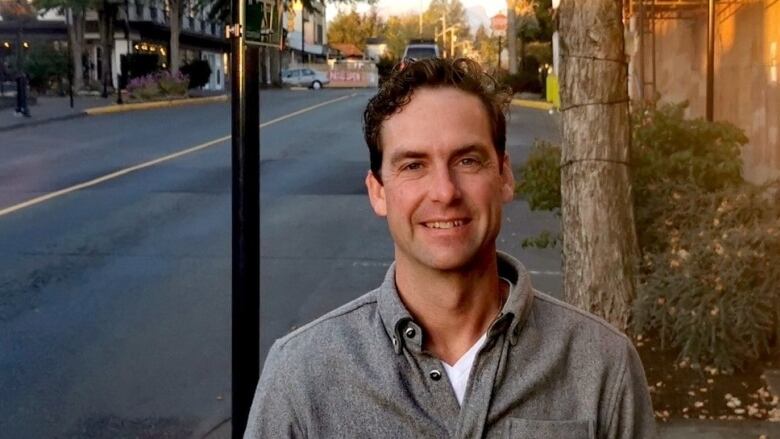 A man in a grey button-up shirt stands on a city sidewalk.