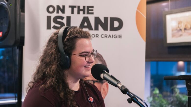 A woman with headphones speaks into a microphone in front of a banner that says, On the Island.