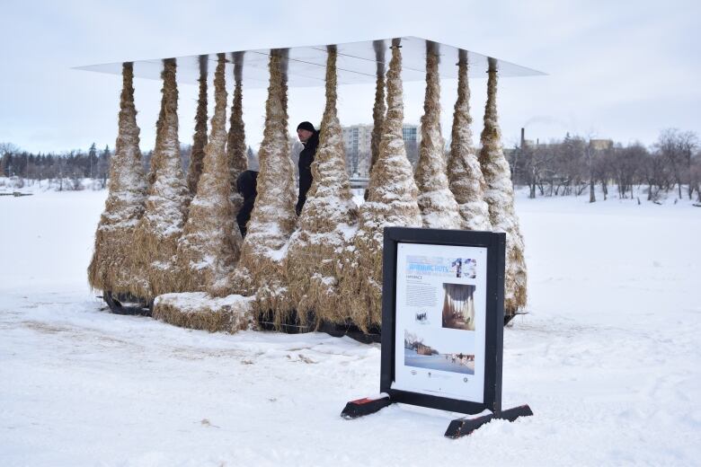 Columns of hay, wide on the bottom and tapering to thinner at top, hold up a reflective cover. The structure is outdoors surrounded by snow.