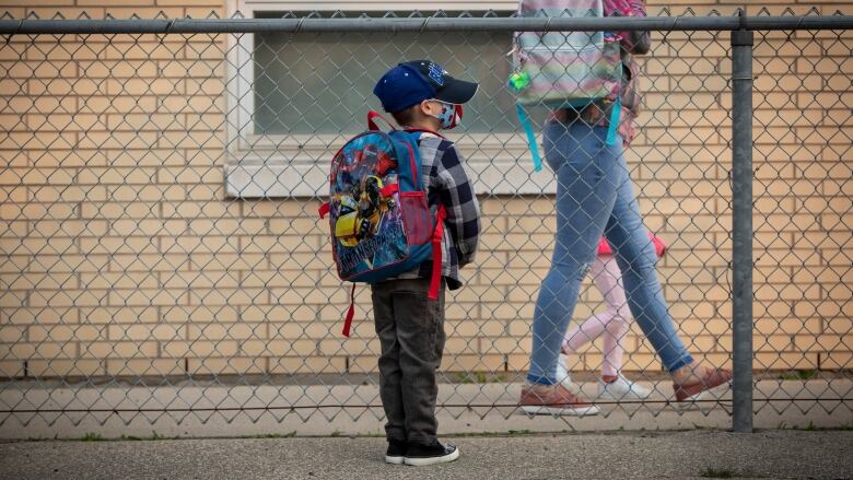 A child wears a hat, face mask and backpack waiting outside a school.