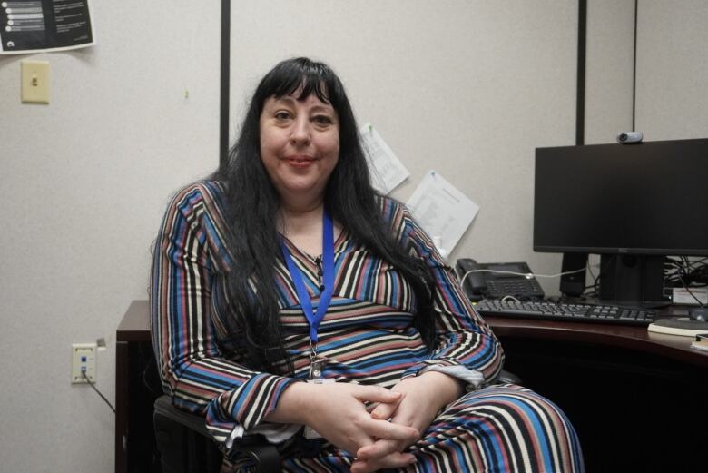 A woman sits at her desk in an office and looks at the camera.