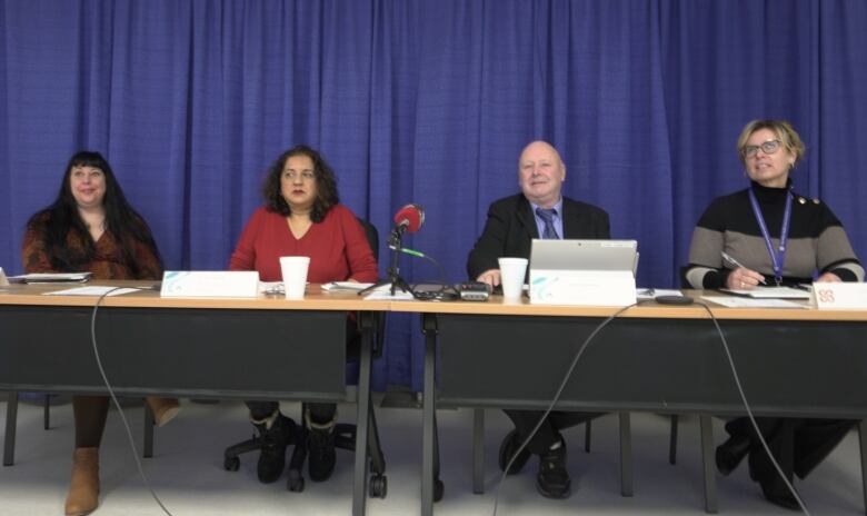 Four people sit in a row behind a table. A microphone sits on the table and a blue curtain hangs behind them.