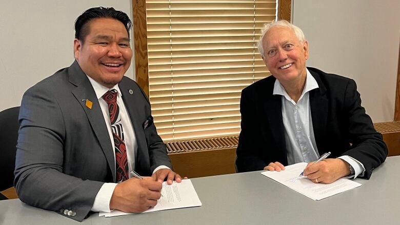 A First Nation Chief with black hair and a grey suit smiles and holds a pen. An older white man representing the B.C. also smiles and holding a pen