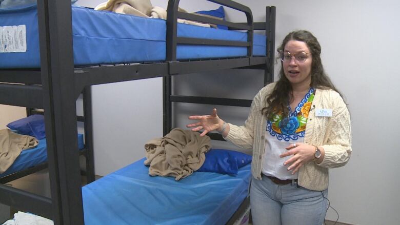 A woman in a cream-coloured sweatshirt gestures next to a bunk bed with blue sheets and pillows.