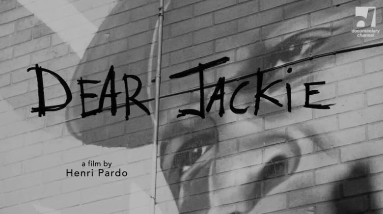 Black and white photo of a mural with the face of a Black man painted on a brick wall. He's wearing a hat. The words in Dear Jackie over the mural in black.