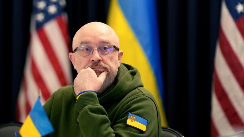 A man wearing glasses and backed by flags rests his head on his chin.