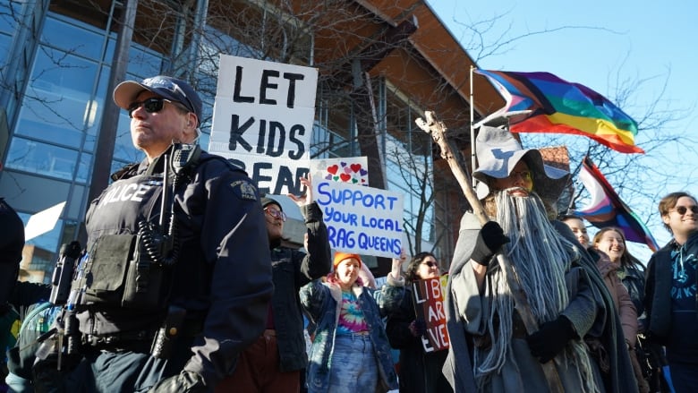 A police officer stands in the foreground while protesters hold banners and wear costumes, such as Gandalf from Lord of the Rings, to support a Drag Queen Story Time in Kelowna B.C.