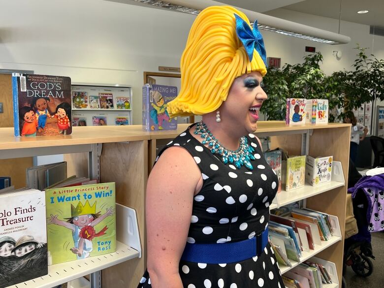 A drag queen with a large yellow wig and white and blue polka-dot dress smiles while looking away from the camera.